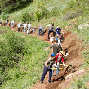 A line of volunteers working on a dirt trail along a green hillside